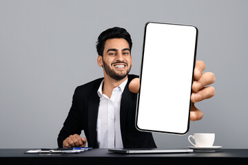 Poster - A young Arab entrepreneur sits at a desk in a modern studio, smiling confidently as he holds up a blank smartphone screen. He is dressed in a formal black suit, exuding a successful lifestyle.