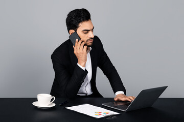 Poster - A Middle Eastern businessman is focused on a phone call while typing on his laptop in a modern studio.