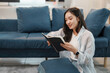 © amnaj - Young woman enjoys a quiet moment at home, sitting on the floor and reading a book by a comfortable sofa in her living room
