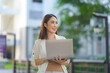 © amnaj - Asian businesswoman smiling and holding laptop while standing in a green urban business district