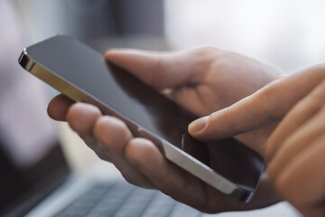 Poster - Closeup view of a man engrossed in his smartphone tasks with an open laptop nearby set against the blurred bustle of an office scene