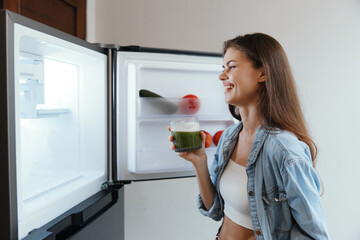 Canvas Print - Young woman smiling while holding a healthy green smoothie, standing in front of an open fridge filled with vegetables, promoting healthy eating and lifestyle