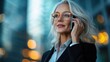© svastix - A stylish woman with gray hair and wearing glasses is speaking on her cellphone against a backdrop of skyscrapers, suggesting a modern and confident lifestyle.