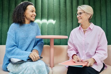 Smiling young multiethnic woman talking to senior female colleague exchanging work experience or assisting coworker with professional development, while sitting in lounge area of coworking space