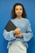 © Seventyfour - Vertical portrait of young multiethnic web designer dressed in elegant sweater holding laptop in hands looking confidently at camera while standing against blue wall