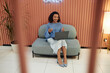 © Seventyfour - Full length shot of cheerful young multiethnic woman presenting during online conference joining video call via laptop, while sitting on comfy couch in lounge area of modern coworking space