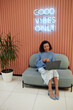© Seventyfour - Vertical shot of smiling multiethnic woman participating in online conference using laptop while sitting on comfy couch in lounge area of modern coworking space