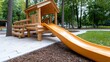 © svastix - A wooden playhouse with an orange slide, surrounded by trees and mulch, offering a fun play area for children in a natural park setting.