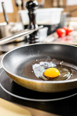 A close-up of a frying pan on a stovetop with a fresh egg cracked open, showcasing the bright yellow yolk and clear egg white. The kitchen background features tomatoes and cooking utensils.