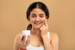 © Prostock-studio - A young Indian woman focuses on her skincare routine, smiling while applying cream to her face. She enjoys a moment of self-care in a simple, isolated setting, emphasizing her beauty and wellness.