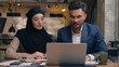 © Yuliia - Two business colleagues co-workers talk discuss corporate work at office cafe multiracial male female employees Indian man muslim woman in hijab diverse businesswoman businessman write tasks laptop