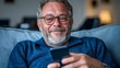 © Radomir Jovanovic - A cheerful man with gray hair and glasses sits on a couch using a smartphone, conveying a sense of relaxation and modern connectivity in a cozy home setting.