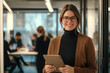 © Myimages - A professional woman in her late thirties, wearing glasses and smiling warmly at the camera as she stands holding an iPad inside a modern office space with people working behind her.