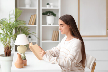 Wall Mural - Beautiful young happy woman reading book at table in living room