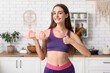 © Pixel-Shot - Sporty young woman with dumbbell showing thumb-up in kitchen. Diet concept