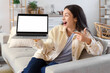 © Pixel-Shot - Beautiful young woman pointing at blank laptop on sofa in kitchen