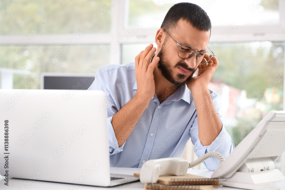 Young businessman suffering from ear pain at table in office