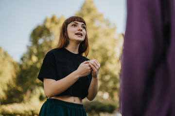 Poster - A young woman wearing a black top speaking with someone outdoors. She is in a park setting with green trees in the background.