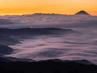 高原の展望台より山を望む、夜明け前の空がオレンジ色に染まり山がシルエット、一面に雲海が広がり山々が半島や島の様に見えて正しく雲海です、外海が荒れて、内海は穏やかです、右上に山、中央に半島で一面が雲海の横構図です。