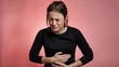 ©  Mohammad Xte - Young Woman in Black Long-Sleeved Shirt, Clutching Abdomen with Pained Expression, Against Soft Pink Background, Conveying Discomfort and Focused Emotion