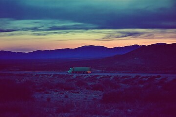 Fototapete - A truck travels along a road at dusk, surrounded by mountains and a colorful sky.