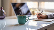 © ahmed elmaslamani - Steaming cup of coffee on office desk with laptop and person writing notes