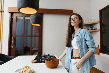 Canvas Print - Young woman in denim outfit smiling while preparing a healthy salad in a bright kitchen with warm tones, promoting healthy eating and lifestyle choices