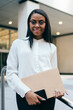 © BullRun - Half length portrait of prosperous black skinned employee in formal wear smiling at camera while holding folder and smartphone.Successful african american businesswoman standing near office building