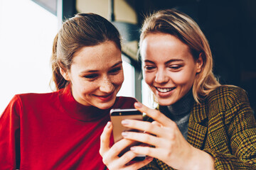Fototapete - Positive female students watching live stream in blog on smartphone connected to 4G internet during exam preparation in cafe.Happy young women reading funny comments in social networks on telephone