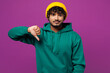 © ViDi Studio - Young sad disappointed dissatisfied Indian man he wear green hoody yellow hat casual clothes showing thumb down dislike gesture isolated on plain purple background studio portrait. Lifestyle concept.
