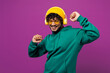 © ViDi Studio - Young happy Indian man wear green hoody yellow hat sunglasses casual clothes listen to music in headphones raise up hands dance isolated on plain purple background studio portrait. Lifestyle concept.