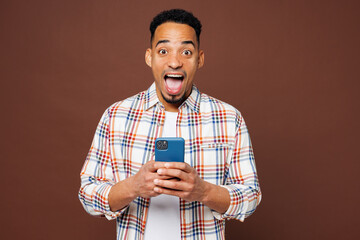 Canvas Print - Young shocked surprised man of African American ethnicity he wears blue shirt casual clothes hold in hand use mobile cell phone isolated on plain brown background studio portrait. Lifestyle concept.