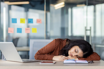 Poster - A young woman appears asleep at her office desk, resting on an open notebook beside a laptop and smartphone, illustrating exhaustion or a need for rest amidst a busy workplace setting.