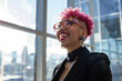 © Sophie - Happy gay businesswoman with pink hair looking out of office city skyscraper window. LGBTQ inclusive deib workplace. Mixed race lesbian executive assistant smiling at work