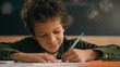 © Yuliia - Little African American guy boy writing notes task class lesson primary school write in copybook kid schoolboy son child pupil student learning listen teacher studying education sitting at table desk