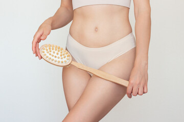 Young Caucasian woman in beige underwear holding a wooden brush with a long handle for anti-cellulite body massage. White background