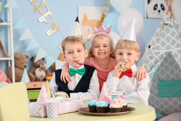 Wall Mural - Little happy kids with cupcakes and gift box blowing whistles while celebrating birthday at table in children's room