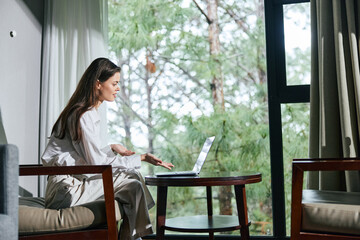 Canvas Print - Young woman in casual white outfit working on laptop at home surrounded by greenery, expressing focus and calmness