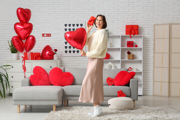 Wall Mural - Beautiful young woman with gift box and air balloon in shape of heart in living room. Valentine's Day celebration