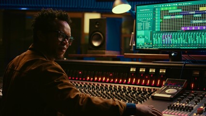 Poster - Portrait of african american audio technician uses mixing console with knobs and sliders in control room, editing recordings and adding sound effects. Skilled music producer at mixer. Camera B.