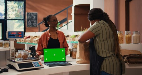Vendor using a laptop with greenscreen layout at store checkout, serving regular customer with natural products. Women chatting next to pc showing isolated display and mockup copyspace. Tripod shot.