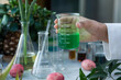 © Nida - Hand holds green liquid beaker, glassware and plants evoke natural laboratory experiment