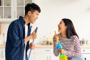 Wall Mural - Funny asian young man and woman in casual singing songs while house-keeping, using dust brush and mop as microphones, white kitchen interior. Domestic lifestyle, house-keeping concept