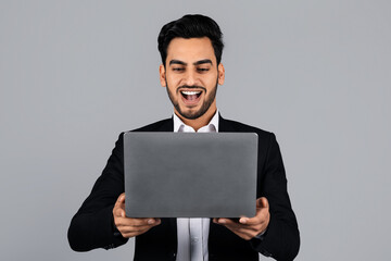 Poster - A Middle Eastern businessman smiles broadly while working on his laptop in a minimalistic studio. Dressed in formal attire, he shows excitement over a recent achievement.