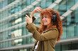 © Cavan Images - Woman with red hair and headphones taking a selfie in a city