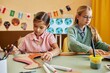 © Seventyfour - Portrait of two young girls painting dinosaur figures made of cardboard while enjoying creative activity in colorful classroom copy space