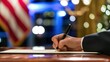 © Vanessa - A hand holding a pen signs a document, with an American flag in the background, suggesting a formal or governmental context.Political career, decision making, leadership, law, administration