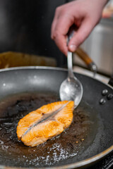 Wall Mural - A close-up of a hand using a spoon to baste a piece of salmon cooking in a hot frying pan, showcasing the sizzling texture and vibrant color of the fish.