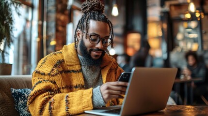 Wall Mural - A man wearing glasses and a yellow jacket is using a laptop and a cell phone