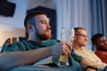 Wall Mural - Side view portrait of bearded young man holding beer glass while watching TV with friends at home and holding beer glass, copy space